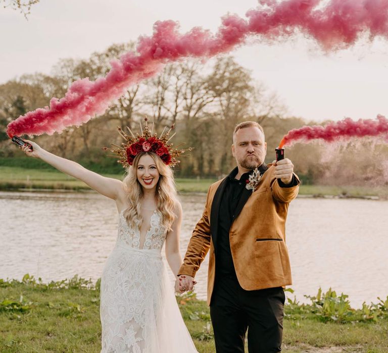 Bride in off shoulder lace wedding dress with corset style top and red rose and gold halo bridal crown holding hands with groom in crushed velvet burnt orange grooms blazer and dried flower boutonniere doing colourful red wedding smoke bomb photoshoot