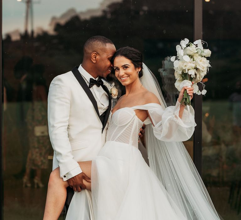 Groom holds leg of bride in white tulle wedding dress and Loeffler Randall wedding shoes while she holds white rose wedding bouquet in the air