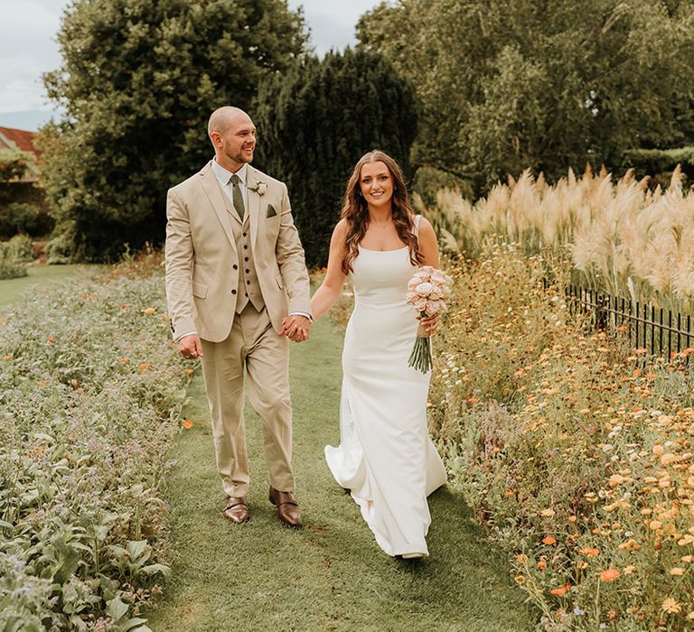 Bride in white slip satin wedding dress walking with the groom in a three piece beige wedding suit 