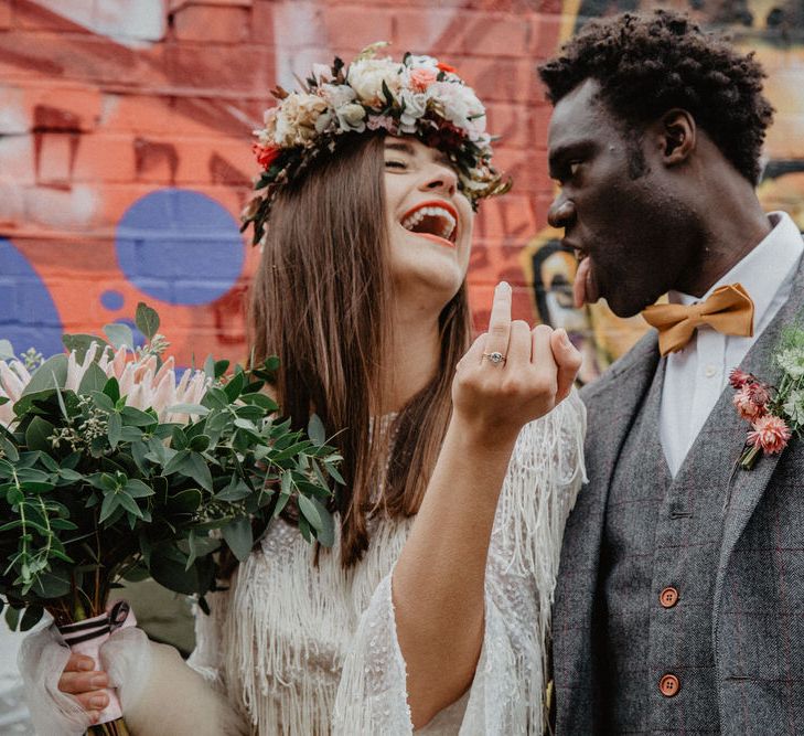 White bride and Black groom both hold up their wedding ring fingers to the camera and make funny faces