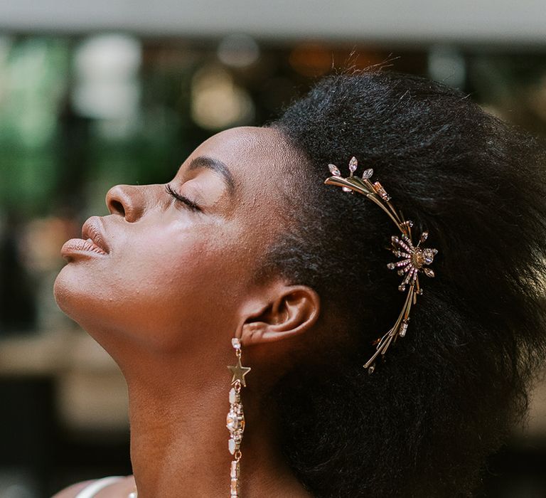 Bride wears her afro swept back with large metallic hair piece to the side. She has flawless dewy makeup and long statement earrings