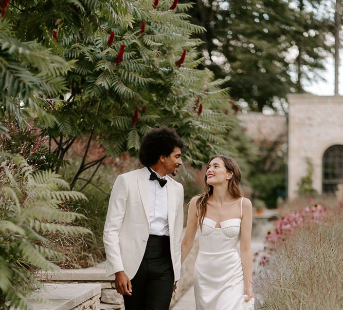 Bride in a satin dress with cup detail and straps holding hands with her groom in a white tuxedo jacket walking through the gardens at Middleton Lodge 