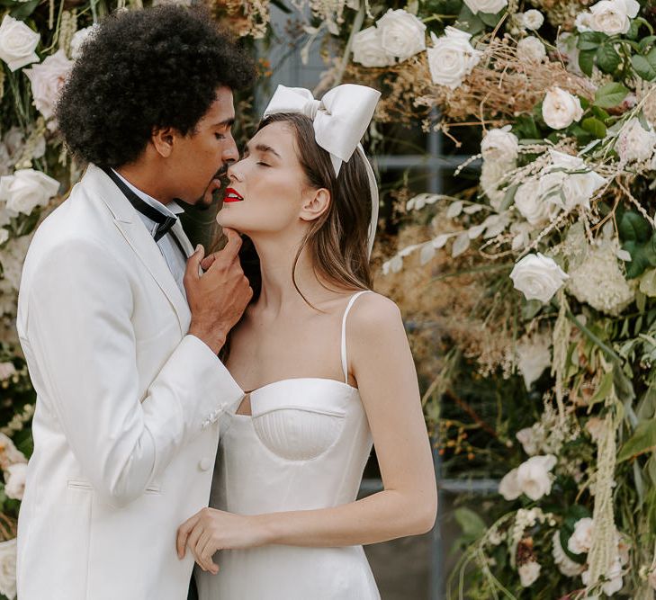 Black groom in white tuxedo jacket kissing his brides nose with long brown hair, red lipstick, bow headdress and satin wedding dress 