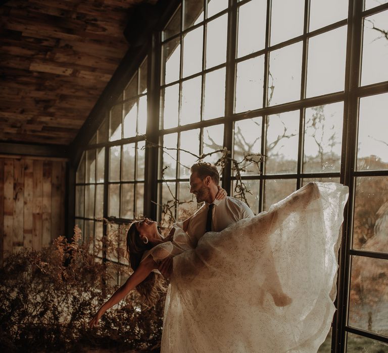 Groom picking up his bride in a sparkly wedding dress in front of the Crittall windows at Hidden River Cabins wedding venue 