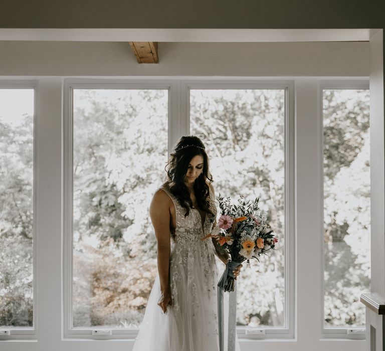 Bride stands in front of window holding rainbow bouquet and wearing BHLDN wedding gown