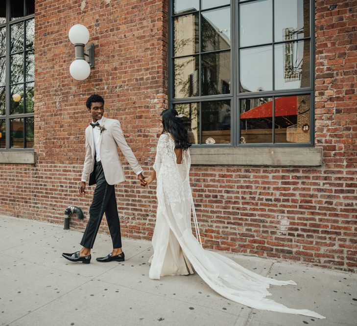 Stylish groom in black tuxedo trousers, white shirt, bow tie, and grey blazer leading his bride in a boho long sleeve mermaid wedding dress 