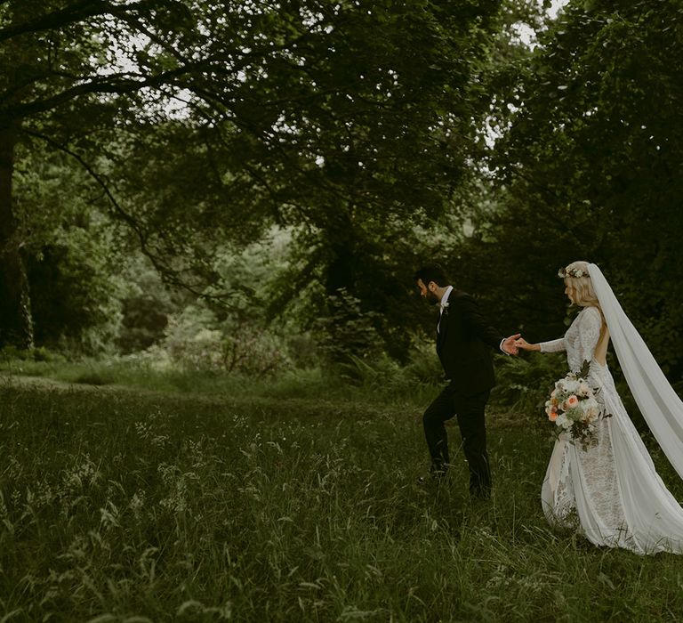 Bride in Grace Loves Lace wedding dress and single tier cathedral length veil walks through forest holding hands with groom in black Hugo Boss suit at Drenagh Estate Wedding