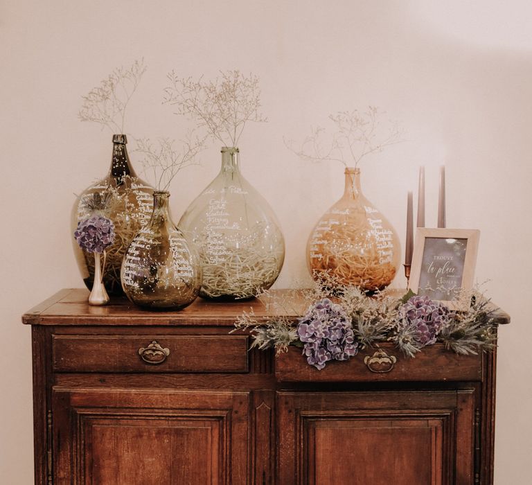 Table seating plan written on glass vases with purple hydrangea flowers