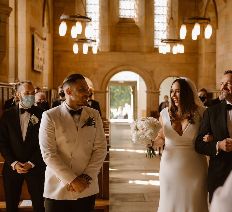 Smiling bride in Made With Love wedding dress stands arm in arm with father in black tuxedo at the church altar next to groom in white double breasted tuxedo