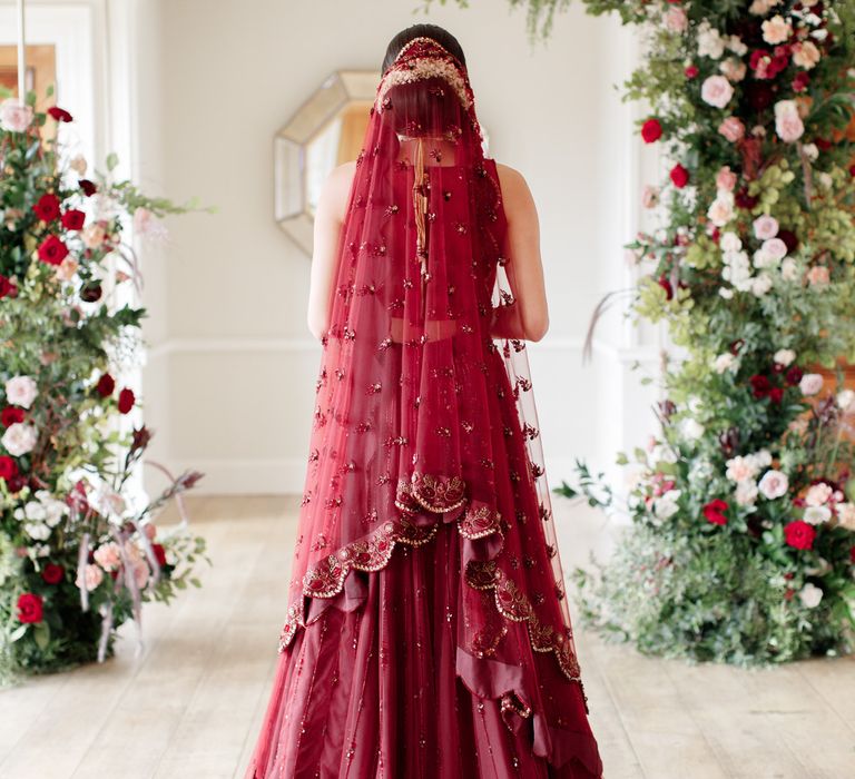 Bride stands with her back to the camera wearing red veil with embroidery 