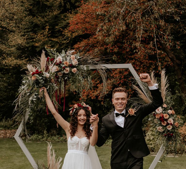 Boho ASOS wedding dress bride with flower crown raising her arms holding hands with her husband in a black tuxedo 