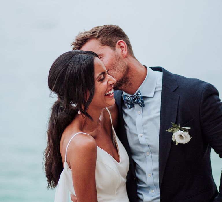 Groom in blue suit with white floral buttonhole kisses bride on the cheek