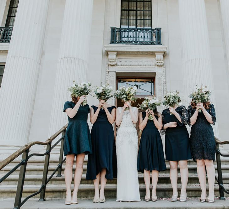 Bridesmaids in front of a town hall all holding bouquets in front of their faces. They are wearing assorted short navy bridesmaid dresses | Joasis Photography