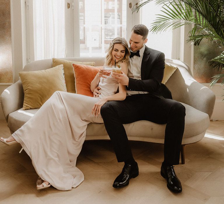 Bride and groom toasting with pink and peach coloured cocktails on a luxe sofa