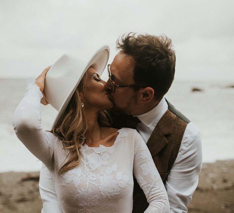 Bride in a fitted lace wedding dress with long sleeves and fedora hat kissing her groom on the beach 