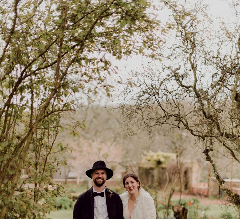 Bride in white Charlie Brear wedding dress with slit and chapel length daisy applique veil stands arm-in-arm with groom in grey tweed suit at garden party wedding in Devon