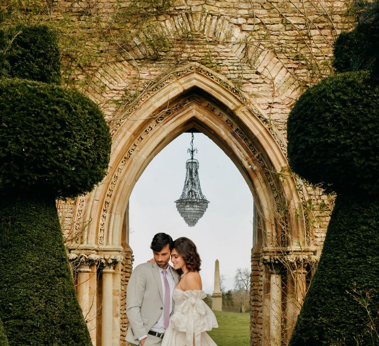 Bride & groom stand on pathway with archway in the background 