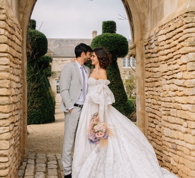 Bride & groom stand beneath brick archway whilst bride wears white full gown