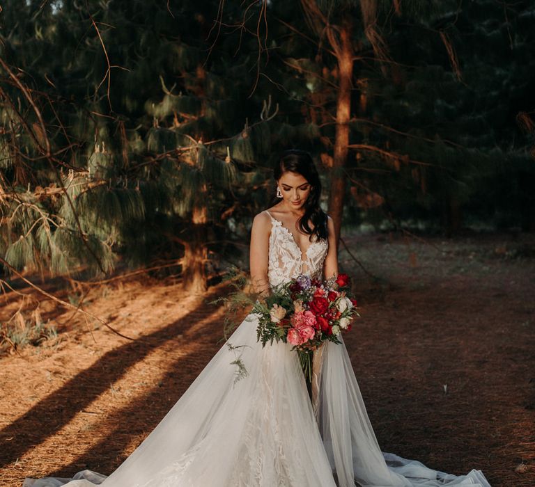 Bride wearing a lace embroidered wedding dress with tulle overskirt, carrying a bouquet of red and pink peonies and roses