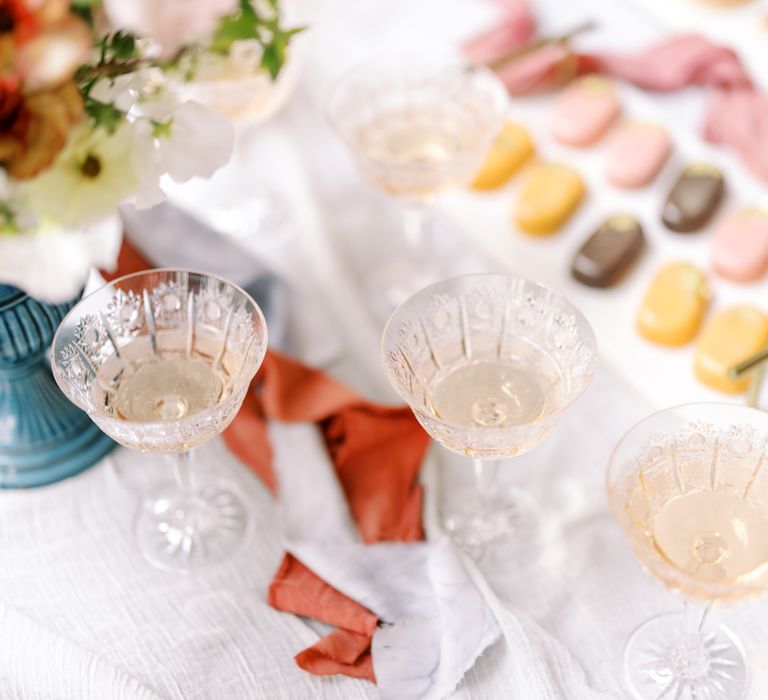 Three glasses of champagne sit on white tablecloth next to pastel coloured biscuits 