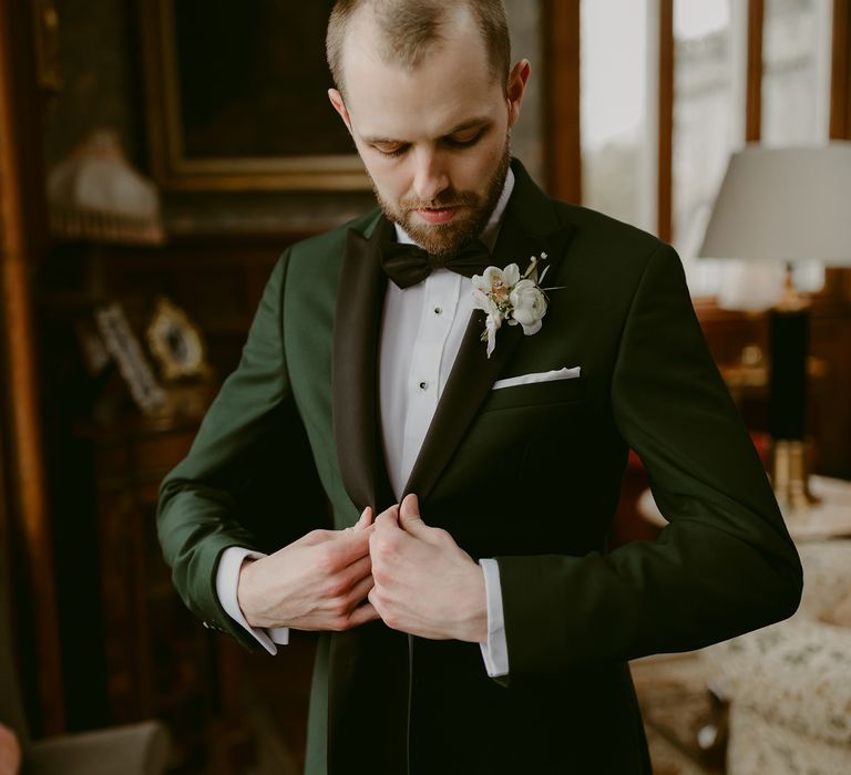 Groom gets ready on the morning of his wedding as he buttons green jacket with black trimming and floral buttonhole