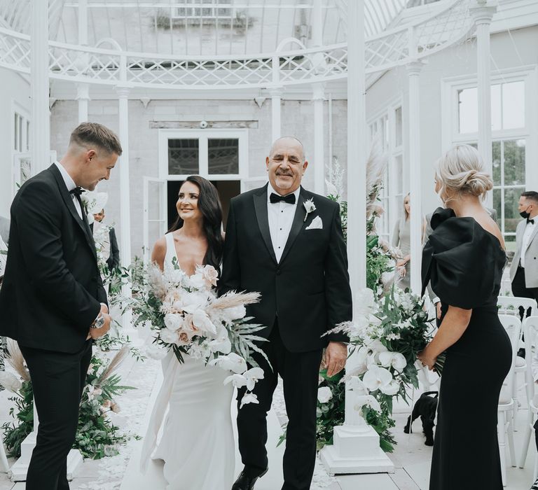 Groom smiles as bride in white Pronovias gown holding white, pink and green wedding bouquet walks down the aisle in the glass conservatory at Came House Dorset arm in arm with man in tuxedo