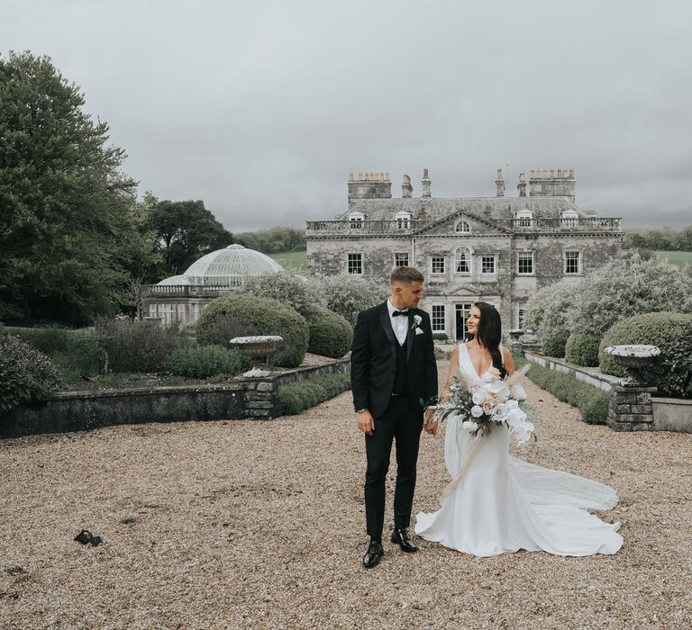 Bride in white Pronovias wedding dress with train holding white, pink and green wedding bouquet smiles at groom in black tux as they stand in the grounds in front of Came House Dorset after their wedding