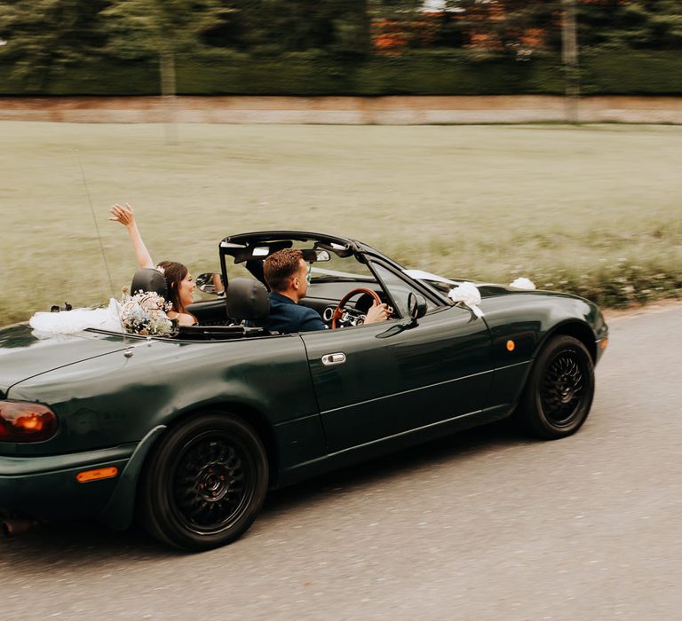 Bride throws arm in the air as groom in blue suit drives green Mazda away from church wedding ceremony