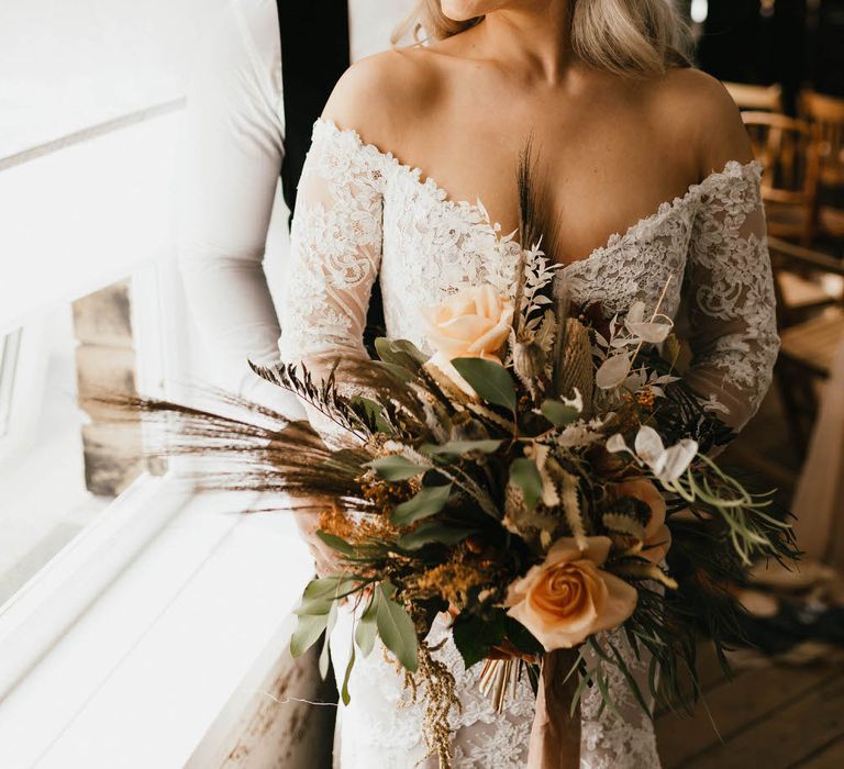 Bride and groom standing by a bright window, looking out. The bride is holding a large bouquet of peach roses and wild grasses