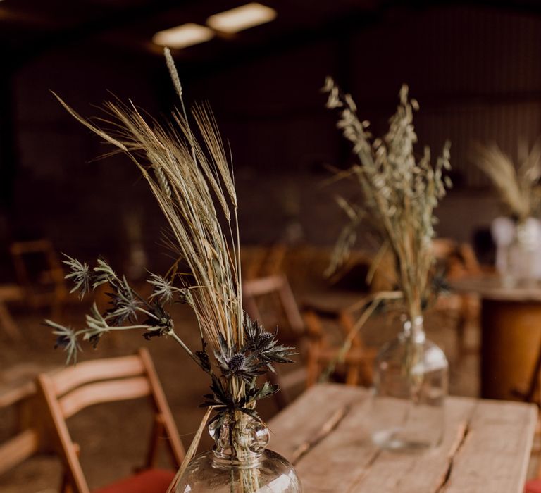 Dried grass in glass jugs on wooden tables inside barn at home farm wedding