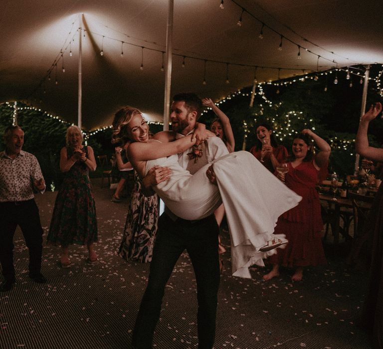 Groom in white shirt and light brown waistcoat swings around bride in white Rime Arodaky wedding dress in fairy lit tipi at garden wedding with burnt orange wedding theme