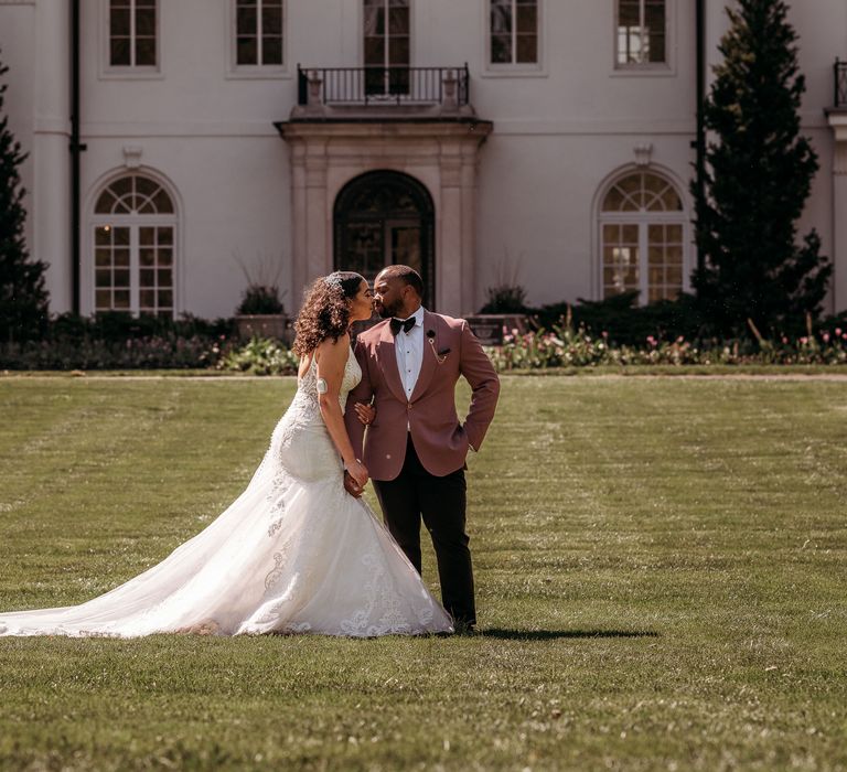 Bride & groom stand with one another in front of manor house within beautiful green gardens