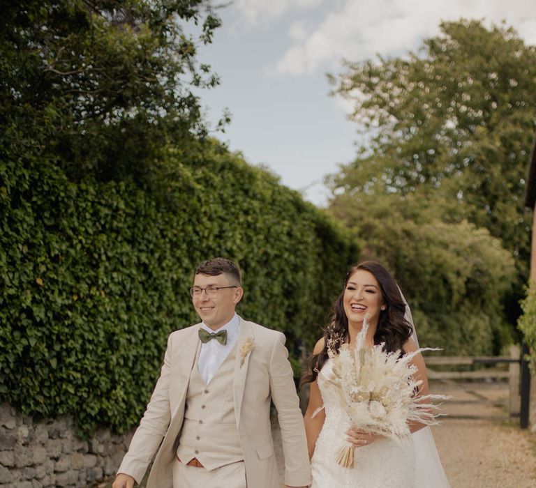 Groom in a three-piece beige suit with sage green bow tie holding hands with his bride in a fishtail wedding dress