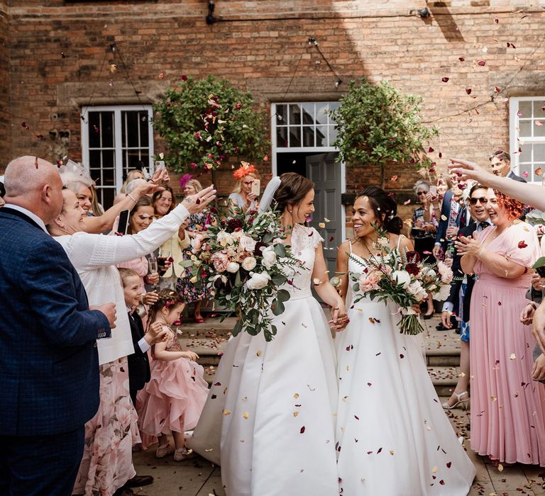 Smiling bride in white cami wedding dress holds hands with bride in white lace capped sleeve wedding dress as they both carry white, pink and green bridal bouquets outside The West Mill Derby whilst guests shower them with multicoloured confetti