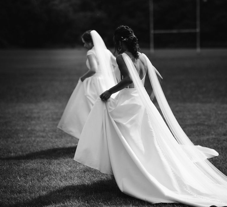 Bride in capped sleeve wedding dress and veil holds up skirts whilst walking across rugby pitch with bride in low back cami wedding dress with shoulder veils after wedding ceremony at The West Mill Derby