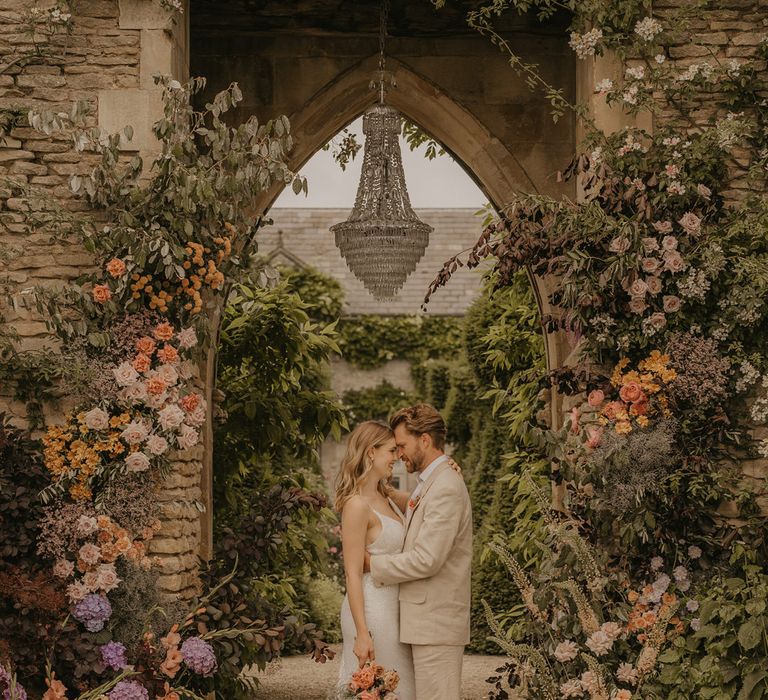 Bride and groom portrait under the castle arch with chandelier at Euridge Manor surrounded by pastel coral, pink, orange and peach wedding flowers 