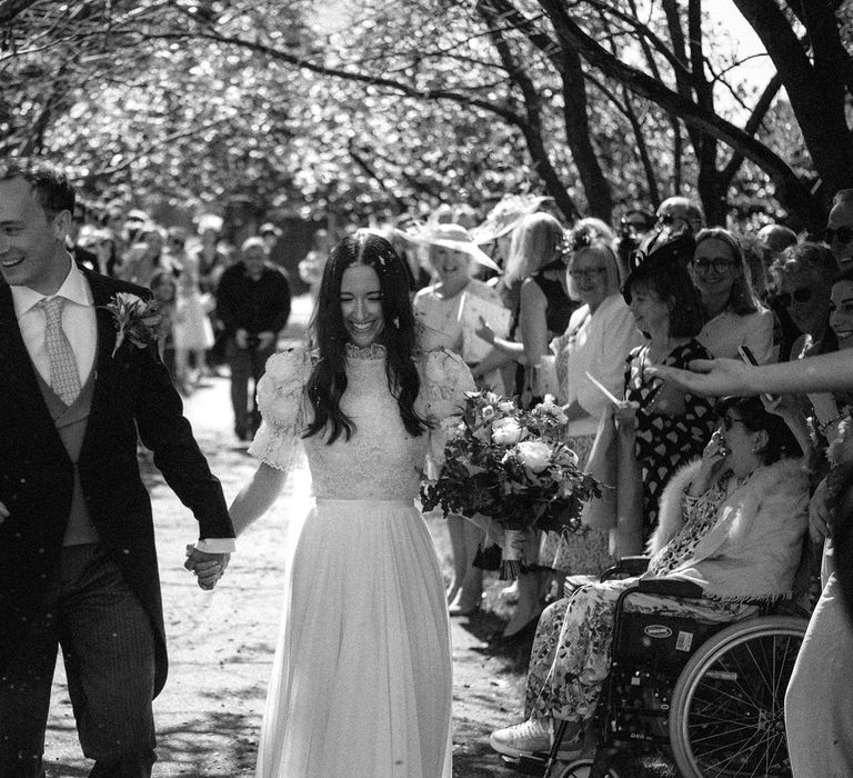 Bride in white lace puffed sleeve Daalarna wedding dress holding white and green bridal bouquet walks down path holding hands with groom in black morning coat as guests throw confetti after church wedding ceremony in Surrey