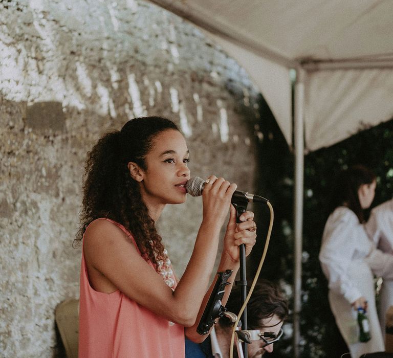  Women in pink sleeveless top and chunky pearl necklace stands singing into microphone at Surrey summer wedding