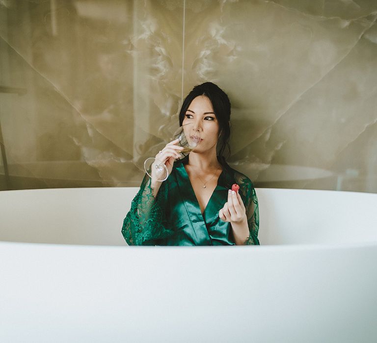 Bride sits in bathtub in green robe whilst eating strawberries and drinking champagne 