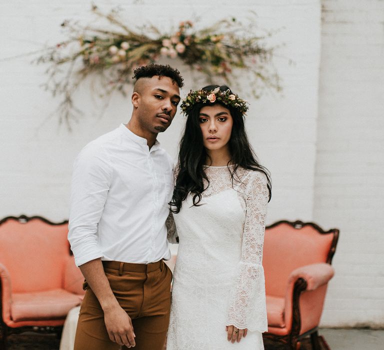 Groom in a white grandad collar shirt and brown trousers embracing his bride in a lace wedding dress with flower crown 