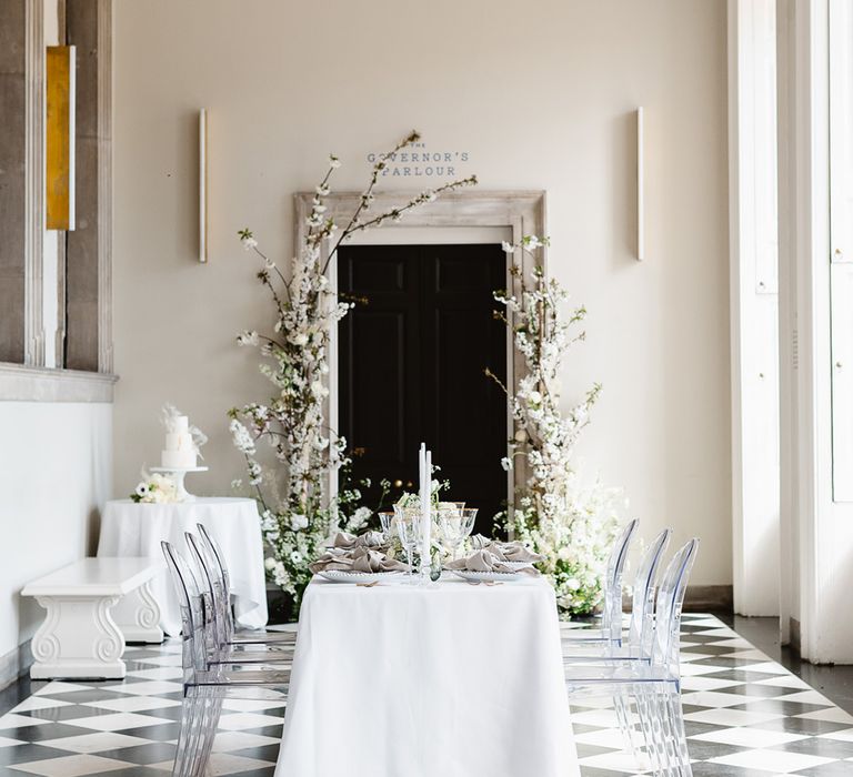 A table with 6 ghost chairs has a white table cloth, white candles and gold rimmed glassware for a black and white wedding reception. 