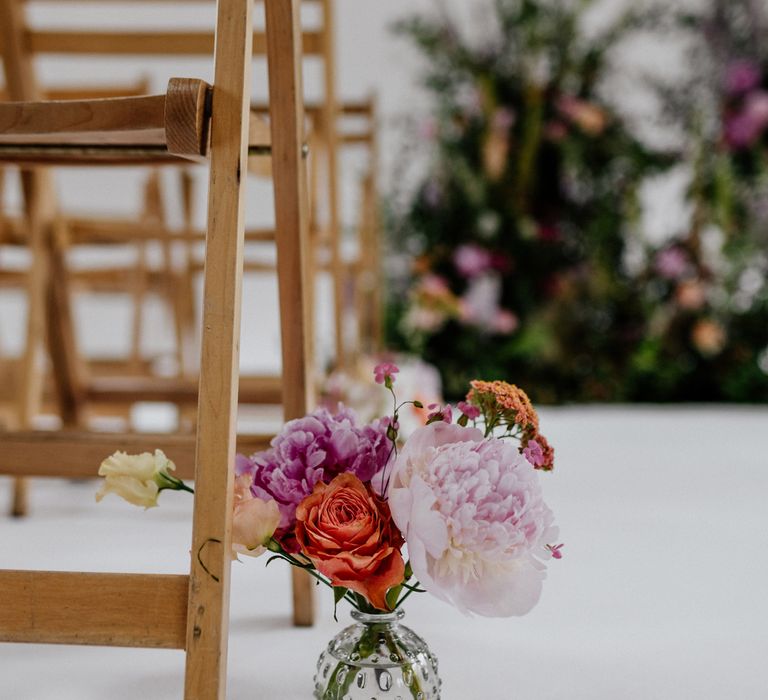 Small glass vase with assorted pink, purple and orange flowers on the floor next to wooden seat in the aisle for wedding at Loft Studios London