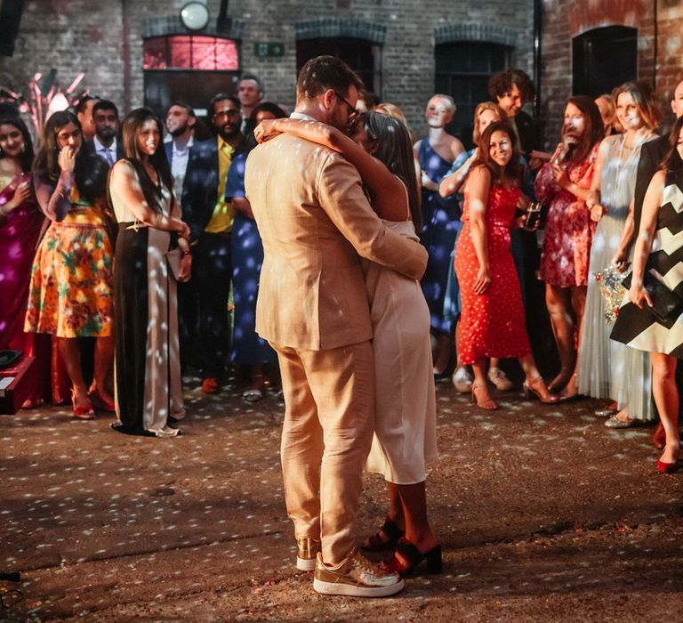 Bride in white satin dress has first dance with groom in linen suit and gold Nike trainers under disco ball whilst guests watch during wedding at Loft Studios London