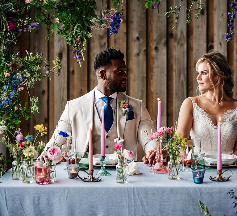 Bride and groom sitting sitting at their sweetheart table decorated with pink taper candles and wildflower stems in vases