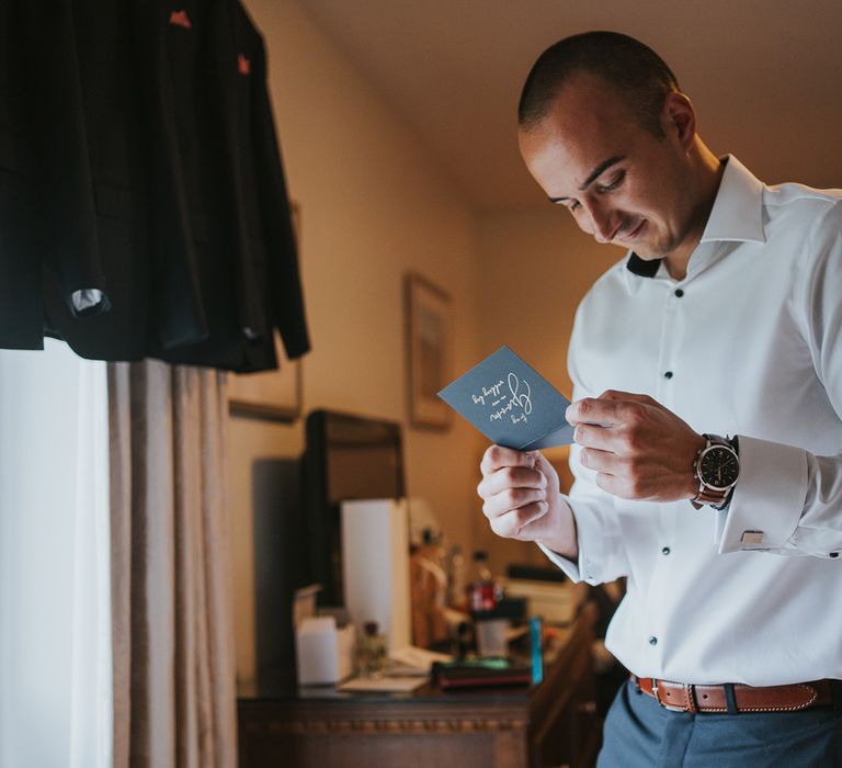 Groom in white shirt and blue suit trousers with brown belt reads message before Tythe Barn wedding with barn wedding flowers