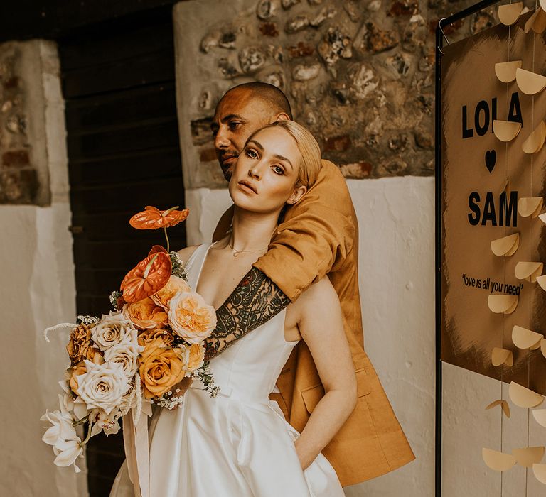 Groom in a mustard suit with tattoos on his arm embracing his bride in a taffeta princess wedding dress with natural makeup and slick back wedding hair 