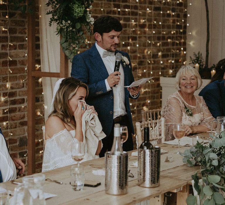 Bride in Grace Loves Lace bardot wedding dress and veil wipes eyes with napkin as groom in blue suit and bow tie makes speech at Isle of Wight wedding with macrame wedding decor