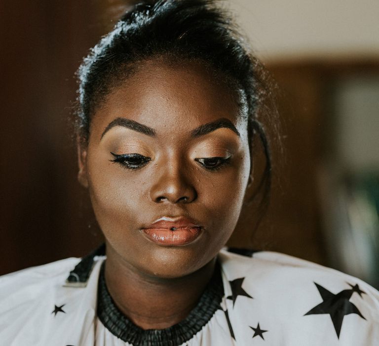 Black bride with defined eye brows, shimmering eyeshadow, winged eyeliner, clear gloss and highlighter on her cheeks having her wedding makeup done