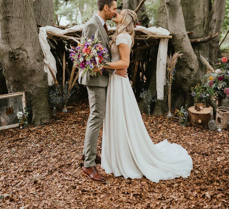 Bride in white lace cap sleeve wedding dress with train holding mixed bridal bouquet kisses groom in grey herringbone suit and brown shoes as they stand in woodland at late summer wedding in Norfolk