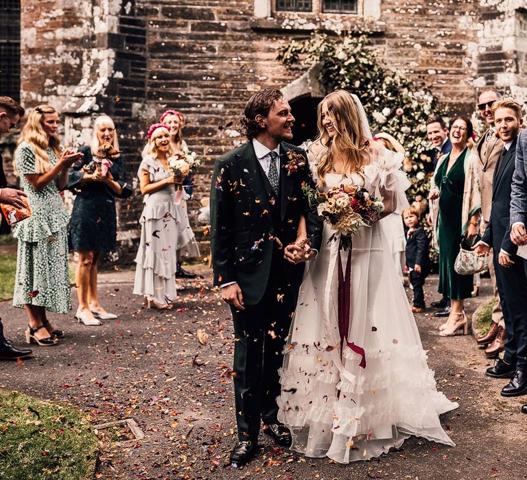 Bride in white Halfpenny London Mayfair dress holding mixed bridal bouquet with burgundy ribbon smiles holding hands with groom in three piece suitas they leave church and guests throw confetti at Cornish wedding